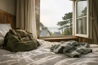 Hotel room with rumpled bed, backpack and jacket near an open window that frames a partial sea view, morning light streaming in, understated and authentic atmosphere.